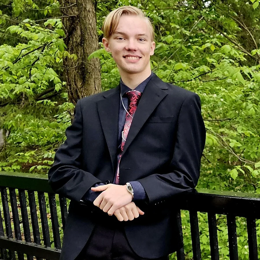 Peter Deems in a dark suit, standing at a railing in front of green trees